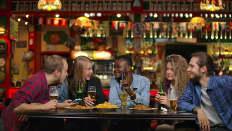 African-American-shows-friends-pictures-on-the-phone-they-look-and-laugh-while-sitting-in-a-bar-in-slow-motion.-Company-of-young-men-and-women.